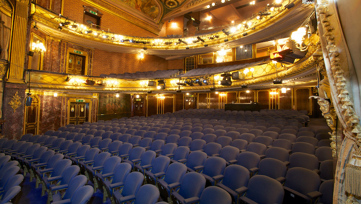 A spacious theater interior with rows of blue upholstered seats facing a stage. The architecture features ornate gold detailing, large chandeliers, and a high ceiling adorned with elegant paintings. The upper balcony is visible, with more seating and decorative elements.