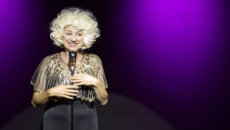 A person with curly white hair stands on stage, smiling while holding a vintage microphone. They are wearing an elegant, ornate top with golden embellishments. The background is illuminated with purple lighting.