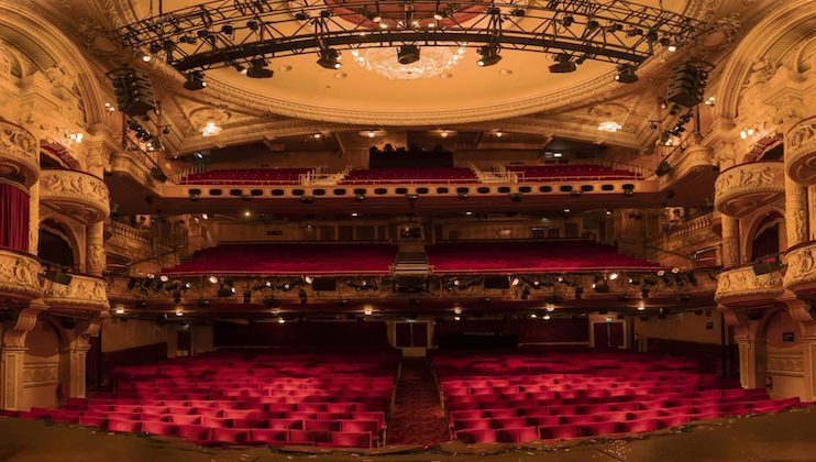 A grand theater interior with ornate designs features multiple levels of seating. The stage view reveals plush red seats, chandeliers hanging from the ceiling, and intricate architectural details on the balconies and walls, all illuminated by warm lighting.