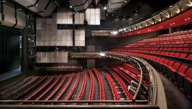 A modern theater with red and black seats arranged in rows facing the stage. The seating includes an upper balcony section. The walls and ceiling are equipped with acoustic panels. The theater is well-lit, and there is no audience present.