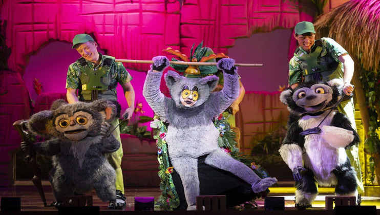 Costumed performers dressed as animated characters from Madagascar on stage. The characters include King Julien, a lemur with a crown, and other animals. The background features a jungle theme with vibrant colors and foliage, and two actors in safari outfits are present.