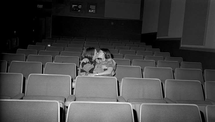 A black-and-white image of two people sitting in an otherwise empty theater. They are embracing and kissing, seated in the middle of the rows of empty seats. The setting is dimly lit with an exit sign visible to the left.