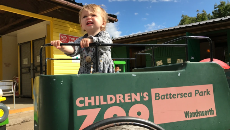 A blonde baby holds the railing of a small vehicle with the branding of Battersea Park Children's Zoo