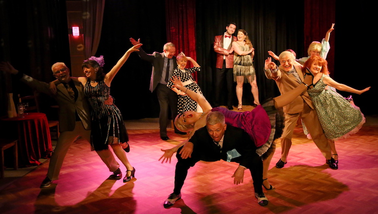 A group of ten people pose in vibrant, theatrical dance positions on a wooden stage with dim lighting. They are dressed in colorful, vintage-style costumes. The scene exudes fun and energy, with couples interacting and performing synchronized movements together.