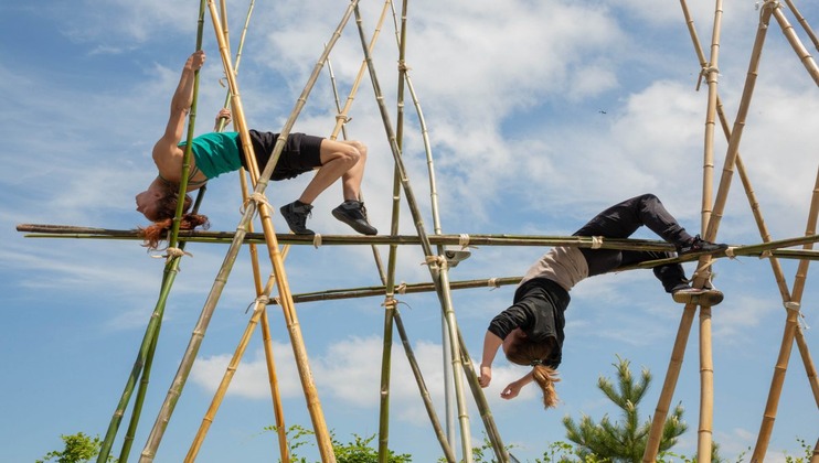 Two individuals perform balancing acts on a bamboo structure against a blue sky with scattered clouds. One person hangs by their arms with their body arched backward, while the other leans backward on the bamboo poles with their arms extended behind them.