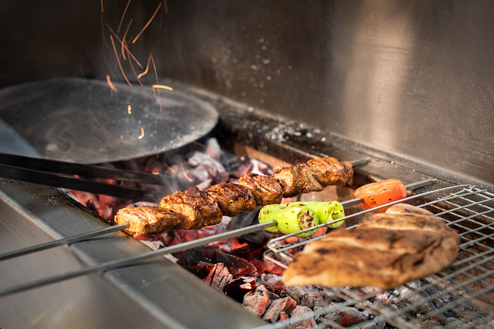 Skewers of grilled meat, a green pepper, and a bread piece are being cooked over a bed of hot coals. A wok pan is visible in the background with sparks flying, creating a lively grilling scene.