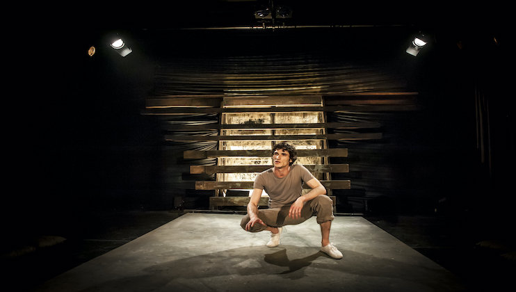 A person in casual attire is squatting on an empty stage, lit by a few spotlights. Wooden slats form a backdrop. The setting is minimalistic and dark, highlighting the person who appears to be in performance.