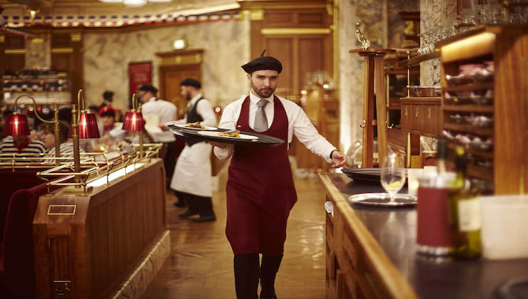 A waiter wearing a beret and maroon apron carries a tray through an elegant, busy restaurant with wooden furnishings. Other staff and diners are visible in the background, and the decor includes marble walls and warm lighting.