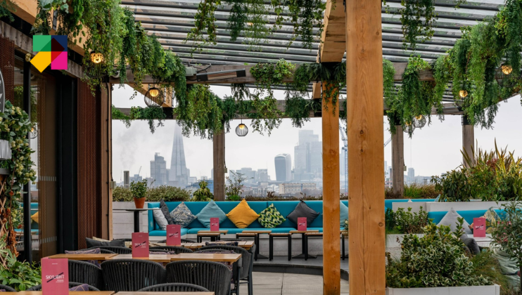 A rooftop terrace features wooden beams adorned with hanging plants and overlooks a cityscape, including modern high-rises and a landmark tower in the distance. The area has tables and chairs, colorful cushions, and is decorated with pink menu cards.