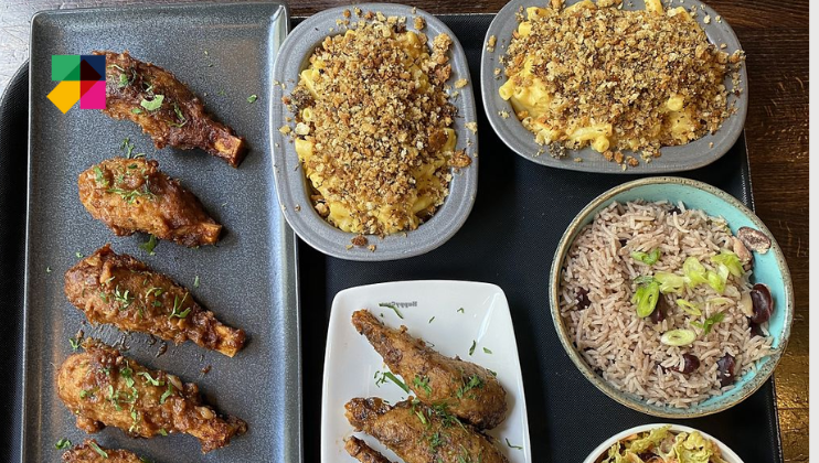 A variety of dishes including crispy chicken wings and drumsticks on a rectangular tray, two bowls of baked mac and cheese with breadcrumbs, a bowl of rice and beans topped with sliced green onions, and a small portion of coleslaw on the side.