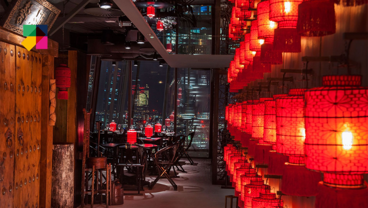 A dimly-lit restaurant with a city view features numerous red lanterns along one wall, casting a warm glow. Dark wooden tables and chairs are set for dining, with additional lanterns on each table. Large floor-to-ceiling windows showcase a nighttime cityscape.