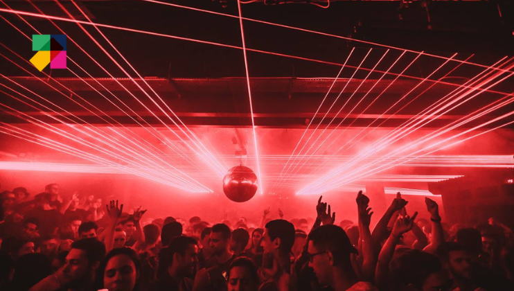 A crowded nightclub with red lighting and laser beams cutting through the foggy atmosphere. A disco ball hangs from the ceiling, reflecting the lights. People are dancing and raising their hands, creating an energetic and lively atmosphere. A logo is in the upper left corner.