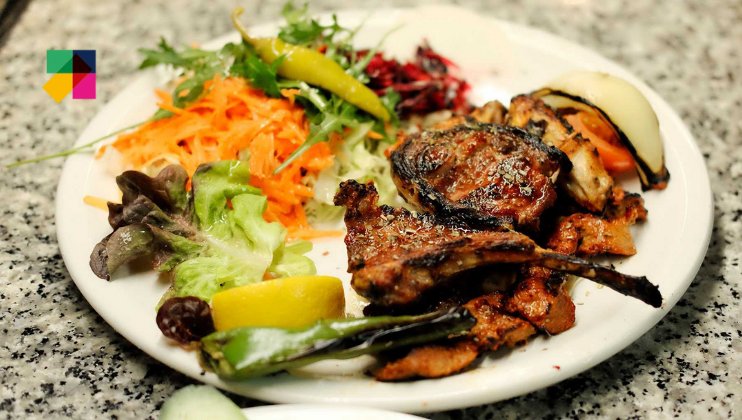 A plate of grilled meat and vegetables, including lamb chops, carrots, greens, peppers, a lemon wedge, and grilled onions. The dish is garnished with herbs and placed on a speckled countertop.