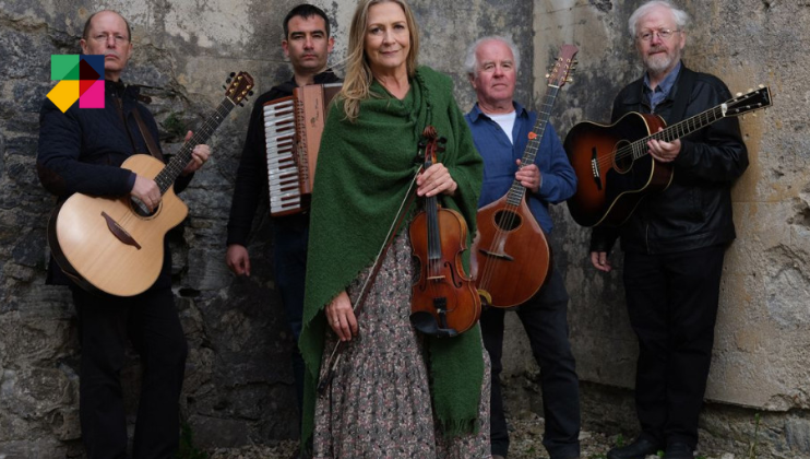 A group of five musicians stands together against a stone wall. They hold various instruments, including a guitar, accordion, violin, and banjo. The central figure, a woman, wears a green shawl and floral dress while holding a violin.