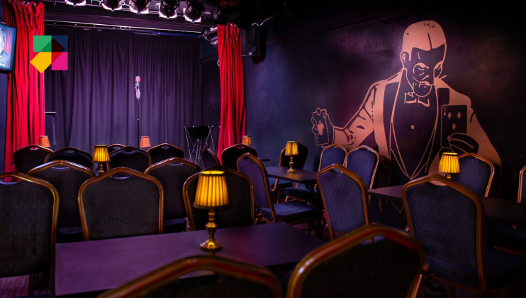 A dimly lit comedy club with a stage at the front, featuring a microphone stand and a black curtain backdrop. Tables with small lamps are arranged in rows facing the stage. The wall to the right has a large artistic mural of a man holding playing cards.