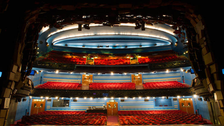 A spacious theater auditorium with multiple levels of red seating, including a ground floor and two balconies. The interior features blue walls and a domed ceiling with modern lighting. The stage area is visible at the front with stage lights overhead.