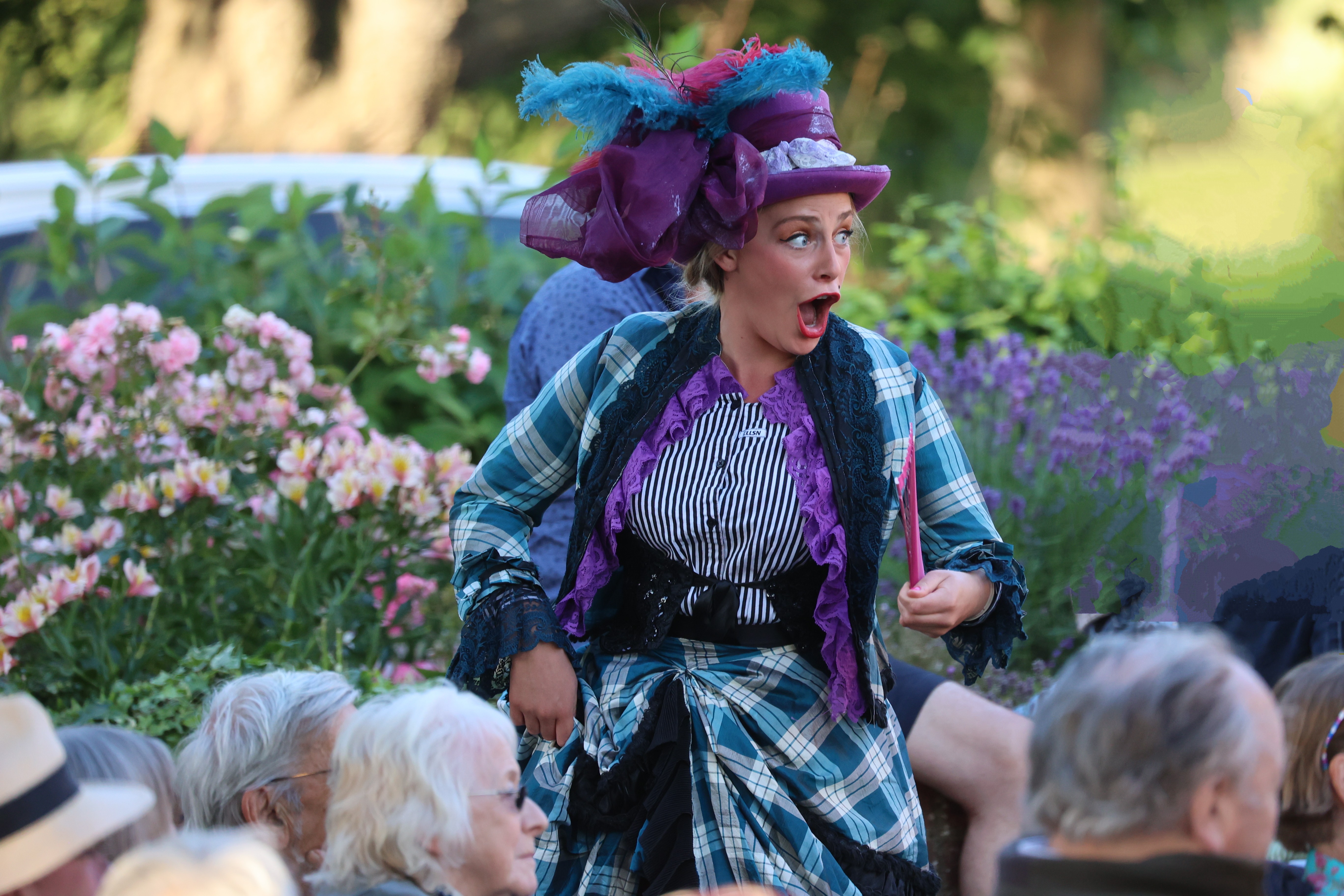 A person dressed in colorful, theatrical attire performs outdoors. They wear a vibrant purple hat adorned with feathers and an elaborate, plaid costume. Their facial expression conveys surprise or excitement. An audience watches among flowers and greenery in the background.
