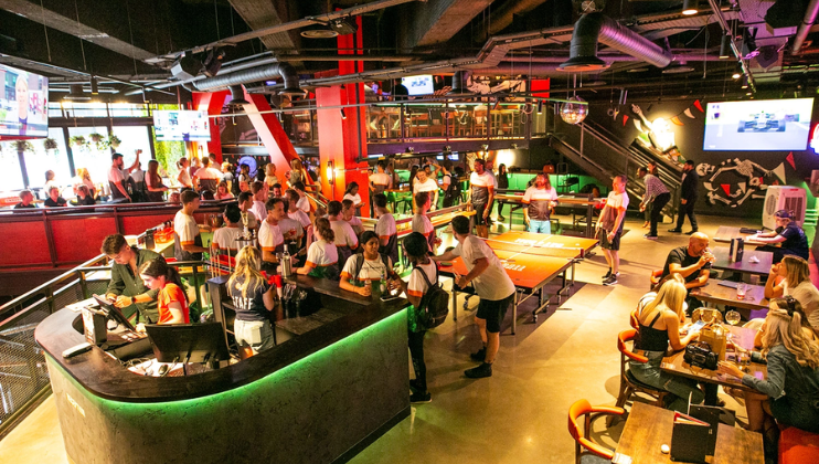 A high shot looking down at the interior at Bat and Ball sports bar in Stratford; in the large space are many tables, and people are drinking, socialising and playing ping pong