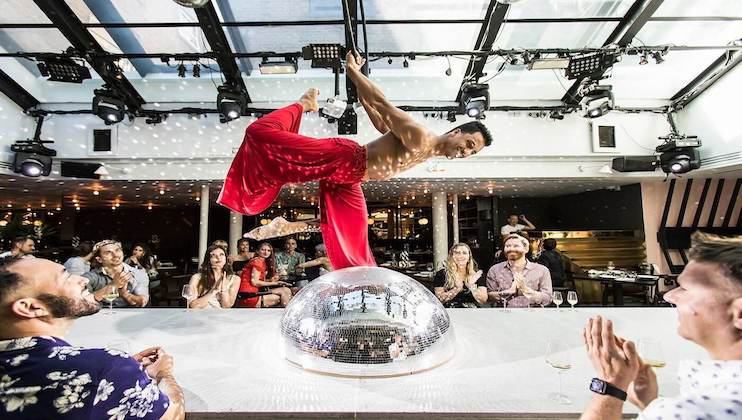 A shirtless performer in red pants balances impressively on a large disco ball inside a well-lit indoor venue. An audience seated at tables nearby watches and applauds, captivated by the performance. The room is adorned with fairy lights and reflective surfaces.