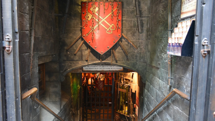 A coat of arms, red, with the symbol of a sword crossing two keys, hangs above the entrance to the Clink Prison Museum; the walls are dark and dingy, and steps lead visitors below ground...