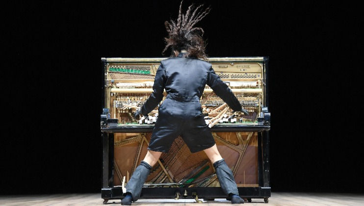 A person with long hair stands in front of an open upright piano with visible inner mechanics, appearing to play energetically. They are wearing a dark outfit and knee-high boots, and their hair is captured mid-motion against a black background.