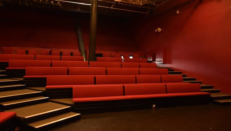 An empty auditorium with tiered seating. The seats have red cushions, and the walls are painted dark red. A black pole is positioned near the center of the seating area. The room is dimly lit, and there is no one present.