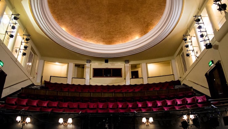 Interior view of an empty theater featuring red seats arranged in rows, a grand stage with a domed ceiling, and elegant lighting fixtures on the walls. The image captures the classic architectural details and serene atmosphere of the space.