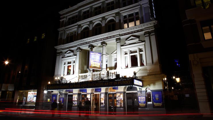 A night view of a well-lit, multi-story theater building with ornate architectural details. The marquee and posters advertise a show, with banners hanging from the upper windows. Blurred taillights suggest passing cars in front of the building.