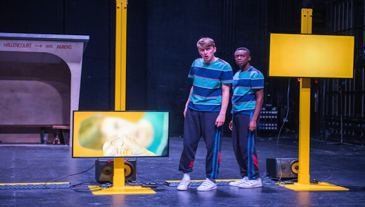 Two actors on stage, both wearing striped blue and black shirts with dark pants and white sneakers. They stand between yellow vertical structures with screens, one showing an image of a child's face. The background is dark and industrial, with minimal set elements.