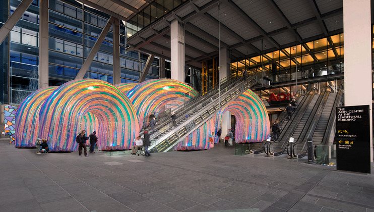 A public art installation features colorful, large, arch-shaped tunnels with a rainbow pattern set outside a modern, glass-fronted building. People walk through the tunnels and use the escalators leading to the entrance.