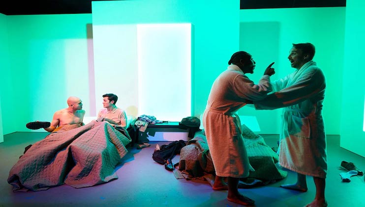 Four people are on a stage with a minimalist set design. Two individuals are seated in bed on the left side, engaged in conversation. To the right, two others stand in bathrobes, one gesturing animatedly towards the other. The background is lit with green and blue hues.