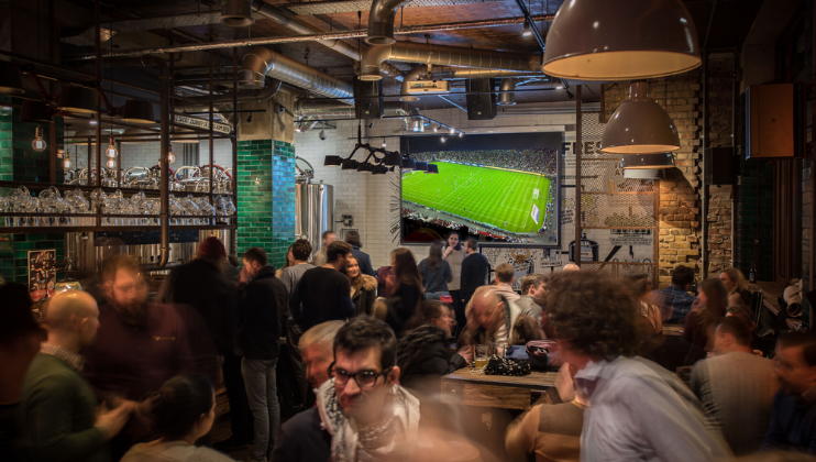 A lively bar with a crowd of people engaging in conversation. A large television screen on the wall displays a soccer match. Industrial decor with hanging lights and exposed pipes creates a cozy atmosphere. Glasses and bottles are visible on shelves behind the bar.