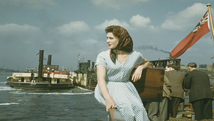 A woman in a striped dress and red scarf sits on the railing of a boat, with a river and a retro steamship in the background. Two men are seen from behind, leaning on the railing. A red flag waves on the right side of the image.