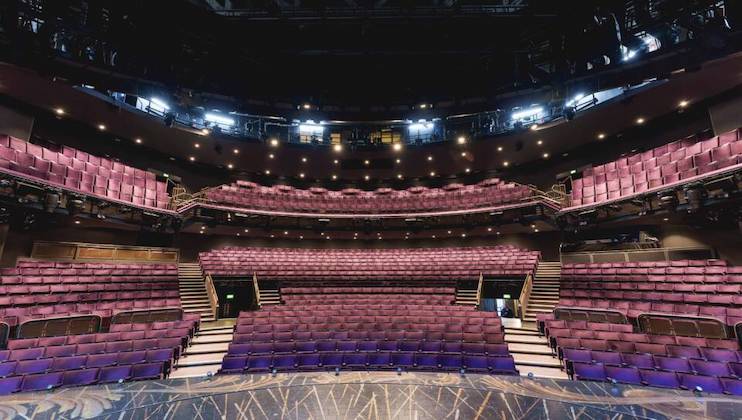 A theater with tiered seating features rows of empty purple seats. The stage floor has intricate designs, and overhead lights cast a moody ambiance. The theater has multiple levels, including a prominent balcony tier. A few people are seen in the distance.