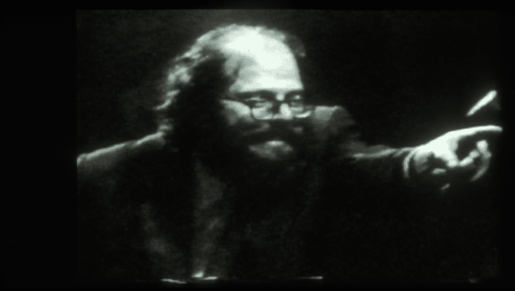 Black and white image of a man with glasses and a beard, pointing emphatically towards something off-screen. His expression is intense, and he appears to be engaged in a passionate speech or performance. The background is dark, adding to the dramatic atmosphere.