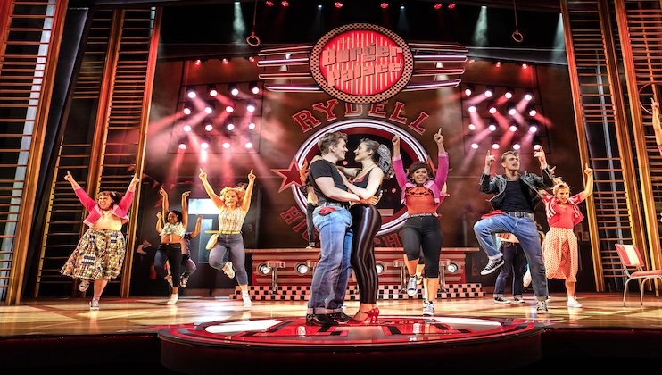 A vibrant stage performance featuring actors dancing energetically. At the center, a couple embraces while other performers strike dynamic poses around them. The stage has a retro aesthetic with a Rydell High sign in the background and bright, colorful lighting.