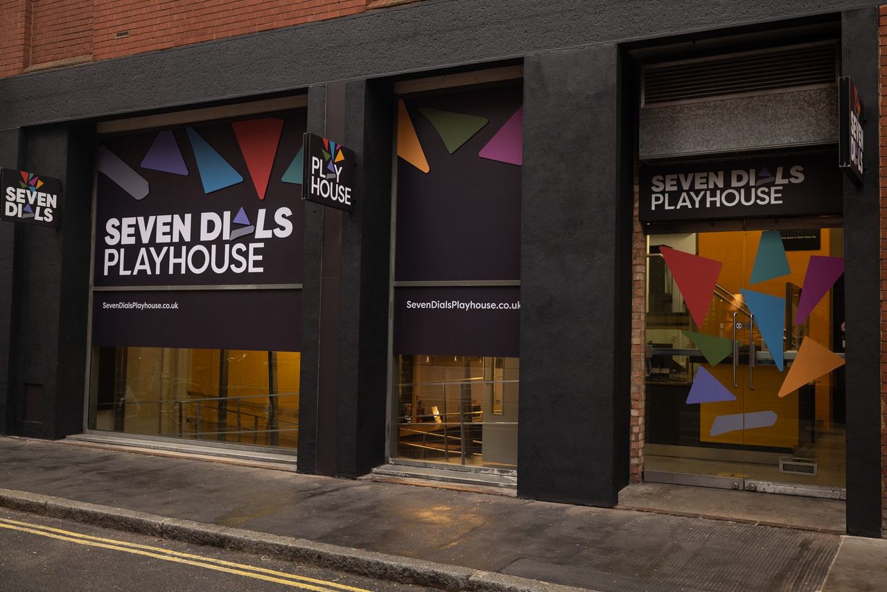 A building facade with large black signs displaying Seven Dials Playhouse in white text and colorful triangular shapes. The entrance has large windows, and a glass door on the right, both reflecting the street and adjacent structures.