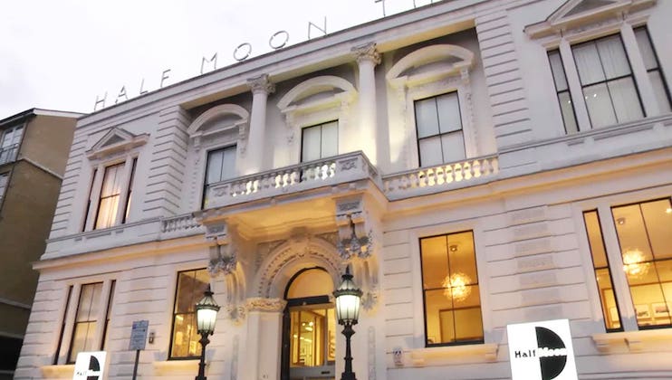 A building with a classical façade, featuring grand arches, tall windows, and ornate details. The sign Half Moon is seen atop the structure. Lit lamps flank the entrance, and warm interior lights shine through the windows. Two signs with the Half Moon logos are visible.