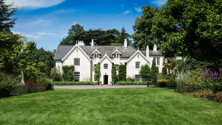 A large, white, two-story country house with dark grey roofs is surrounded by lush greenery. The house has multiple chimneys and windows, and an arched entrance door. The blue sky and well-maintained lawn add to the scenic beauty of the property.