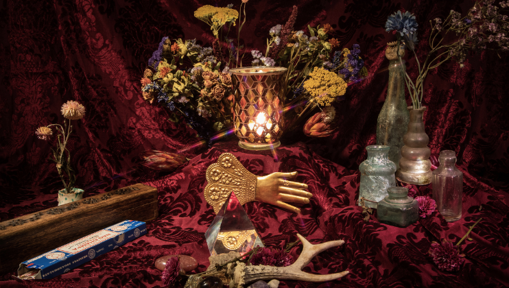 A still life arrangement featuring a flickering candle in a decorative holder surrounded by various dried flowers, vintage bottles, a wooden box, incense, a brass hand ornament, an antler, and other eclectic items on a rich, burgundy fabric background.