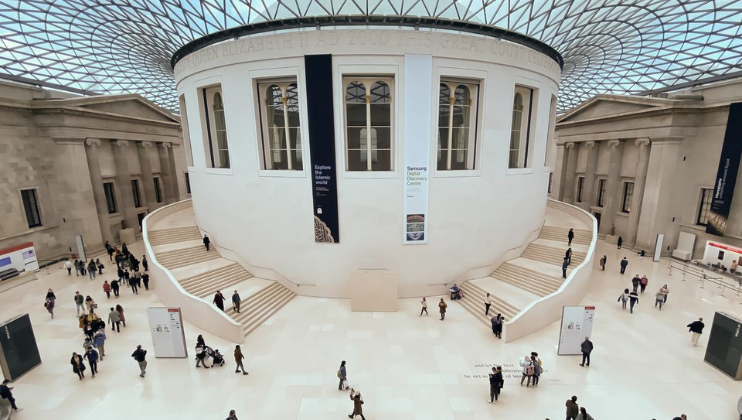 Wide-angle view of the British Museum's Grand Court, featuring a central, round structure with steps leading up to it. People wander around the white marble floor, and the glass roof creates a grid pattern above, flooding the space with natural light.