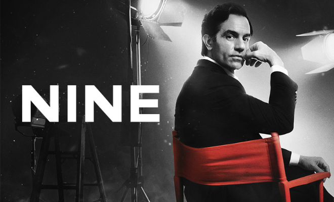 Against a black-and-white background of a movie set, Ramin Karimloo turns to look at the camera from his red director's chair.