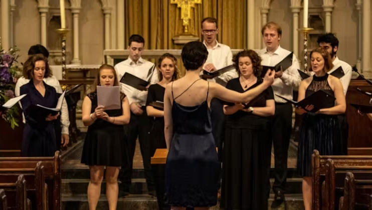 A choir master in a blue dress conducts several singers from Hesperos Choir singing in front of the organ in a beautiful church.