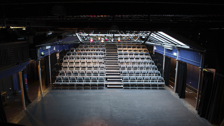 A theater interior with empty tiered seating facing the stage. The seats are arranged in rows, divided into three sections by two staircases. Overhead, several stage lights shine down. The surrounding walls are painted dark blue, and the area is well-lit.