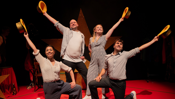 Four performers on stage, each raising one arm high while holding a gold hat. They are standing in a line with joyful expressions and colorful outfits against a dark background with a large star. Two are kneeling in front, and two are standing behind them.