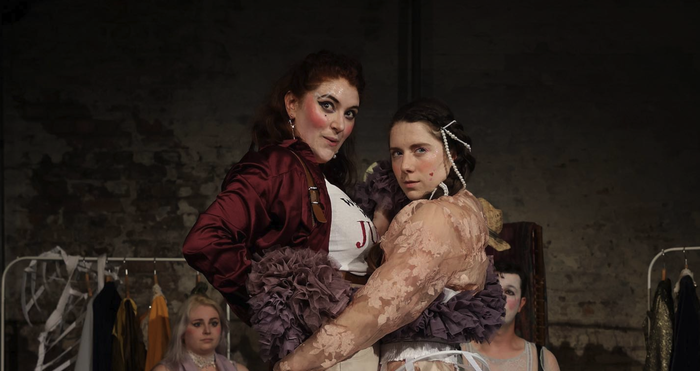 Two performers stand closely, facing the camera with confident expressions. Both sport dramatic makeup and unique costumes; the person on the left wears a maroon jacket and tutu, while the one on the right dons lace attire with ribbons. Other costumed people are visible in the background.