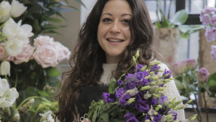 A woman with long dark hair smiles while holding a bouquet of purple flowers. She stands in a floral shop adorned with various flowers in the background, including pink roses and white blossoms. She wears a light-colored sweater and an apron.