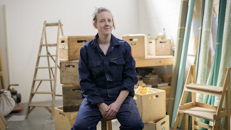 A person wearing a dark blue jumpsuit sits on a stool in a workshop surrounded by wooden crates, a stepladder, and other tools. They have short, light brown hair and a calm expression, looking slightly to the side. The background is brightly lit with natural light.