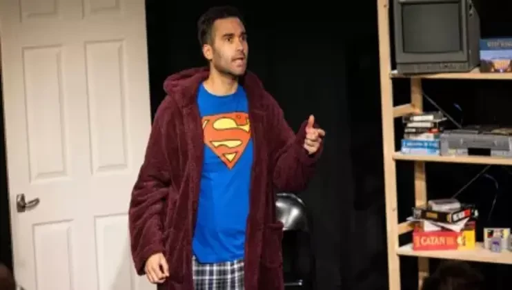 A man stands indoors wearing a maroon robe over a blue Superman t-shirt and plaid pants. He seems to be speaking or performing. Behind him, there is a white door on the left and a shelf with electronics, books, and games on the right.