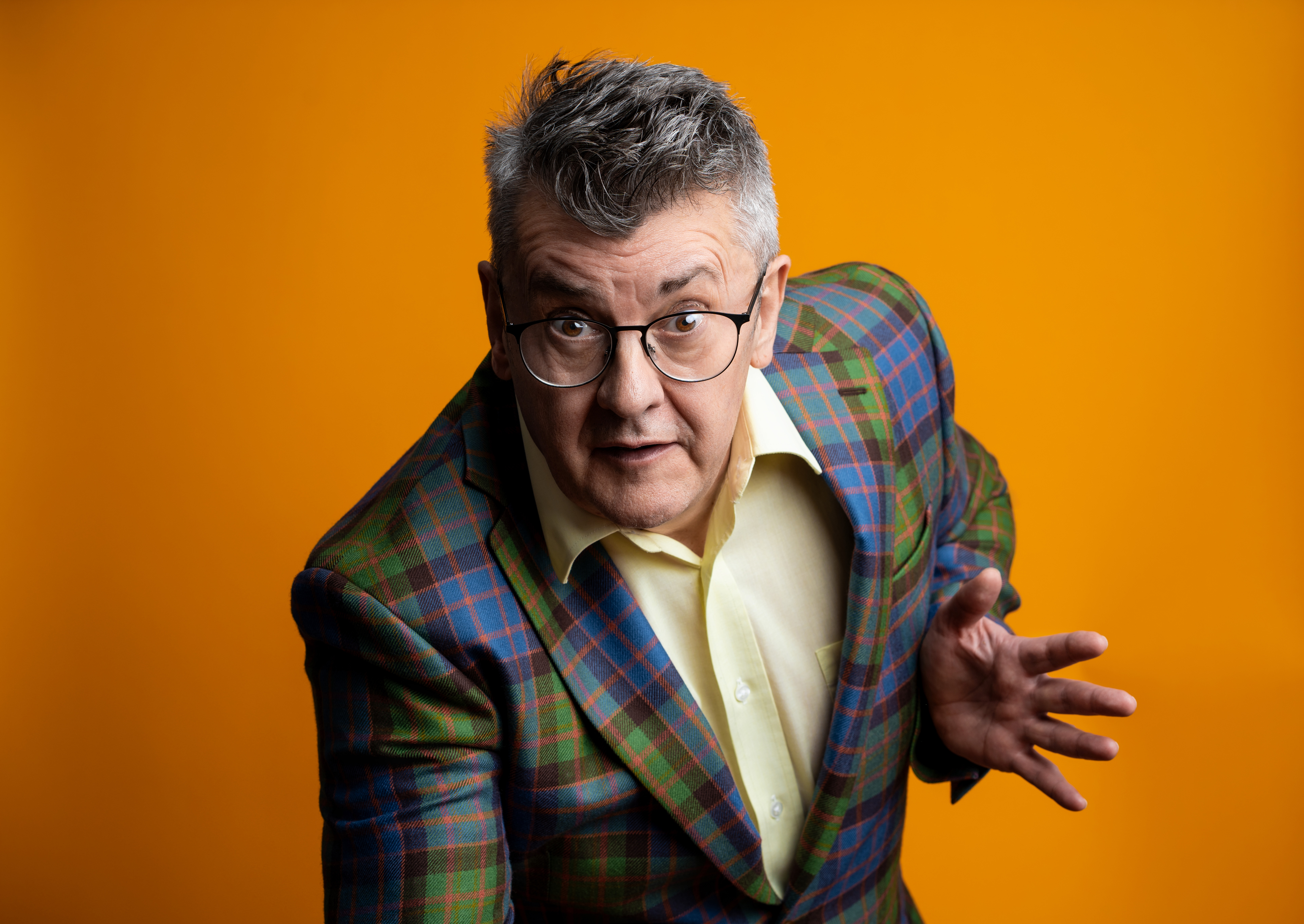 A man with short gray hair and glasses is posed against an orange background. He is wearing a plaid blazer in green, blue, and red, over a yellow shirt, and has an animated expression with one hand raised as if making a point or explaining something.